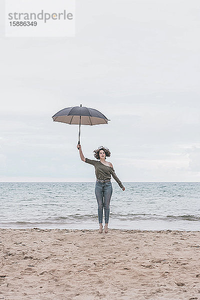 Junge Frau springt am Strand mit einem Regenschirm in der Hand