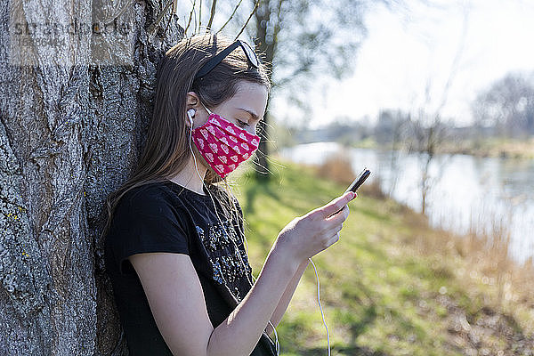 Mädchen mit selbstgemachter Schutzmaske  das ein Smartphone benutzt und sich an einen Baum lehnt