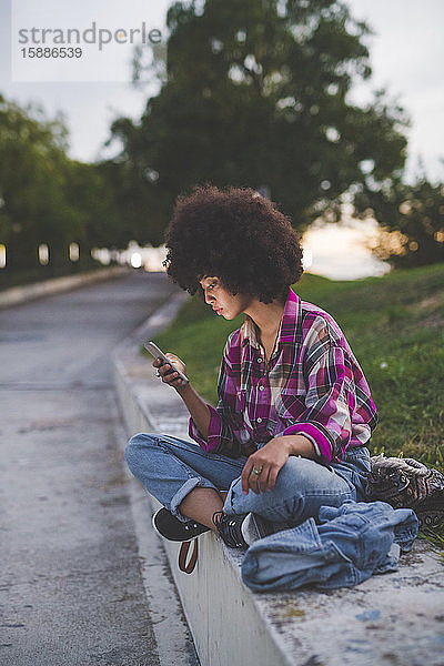 Junge Frau mit Afro-Frisur sitzt mit Smartphone auf dem Bordstein
