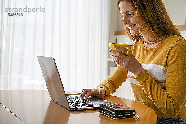 Lächelnde junge Frau bei einer Tasse Kaffee mit Laptop zu Hause