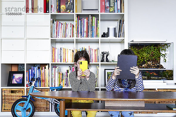 Zwei Schwestern sitzen Seite an Seite am Tisch im Wohnzimmer und benutzen elektronische Geräte