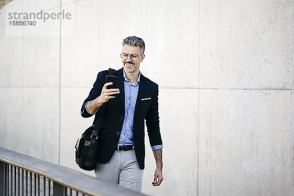 Lächelnder grauhaariger Geschäftsmann geht mit seinem Handy durch die Stadt