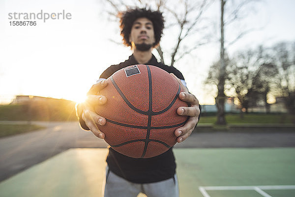 Junger sportlicher Mann posiert bei Sonnenuntergang mit Basketball auf dem Platz