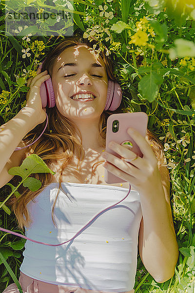Glückliche junge Frau mit Kopfhörer und Smartphone liegt im Frühling auf einer Blumenwiese