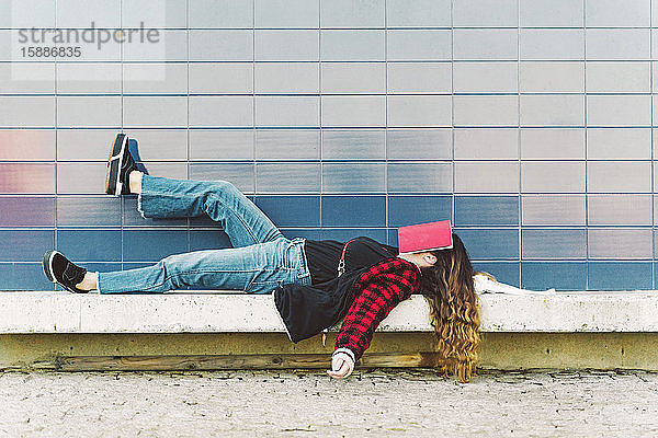 Teenager-Mädchen liegt auf einer Wand im Freien und bedeckt das Gesicht mit einem Buch