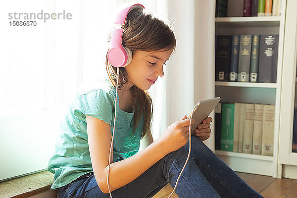 Lächelndes Mädchen sitzt zu Hause mit Kopfhörer und digitalem Tablet auf dem Boden