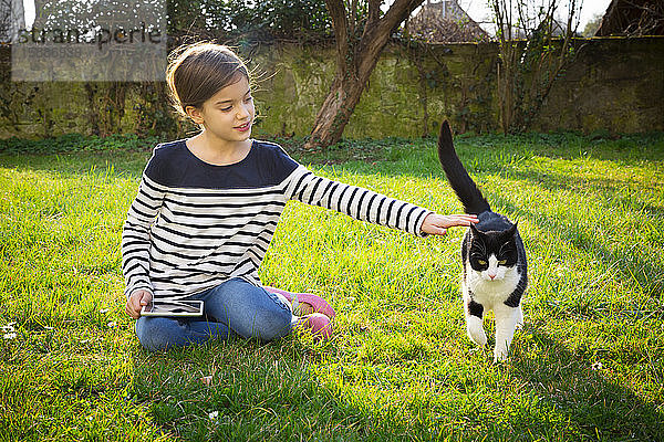 Porträt eines kleinen Mädchens auf einer Wiese sitzend mit Mini-Tablette streichelnder Katze