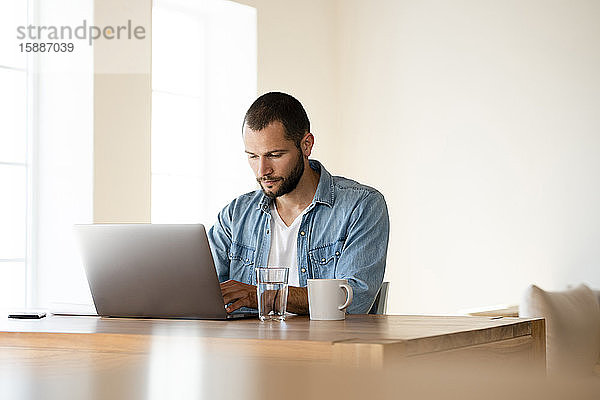 Fokussierter junger Mann arbeitet zu Hause am Laptop im Home-Office im modernen Wohnzimmer