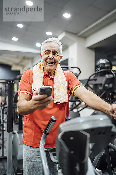 Lächelnder älterer Mann macht Pause und benutzt Handy im Fitnessstudio