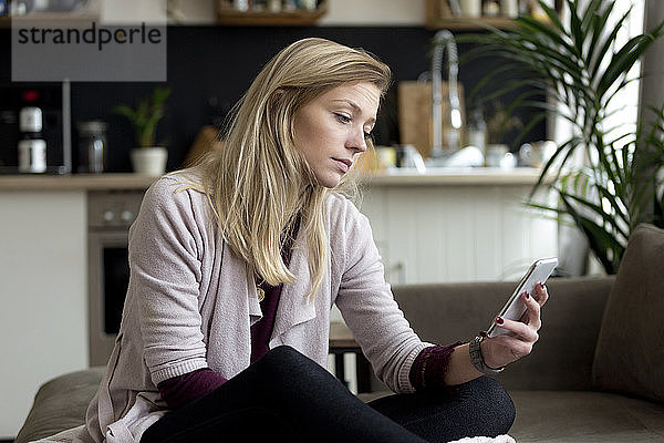 Porträt einer blonden jungen Frau  die zu Hause auf der Couch sitzt und auf ihr Handy schaut