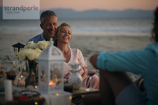 Lächelndes reifes Paar  das einen männlichen Freund ansieht  während es am Strand der Riviera Nayarit  Mexiko  ein Abendessen bei Sonnenuntergang genießt.