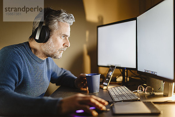 Älterer Mann mit Kopfhörern sitzt zu Hause am Schreibtisch und arbeitet am Computer