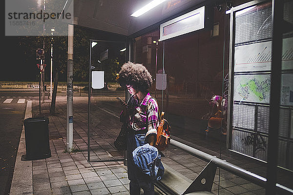 Junge Frau mit Afro-Frisur benutzt Smartphone an Bushaltestelle in der Stadt