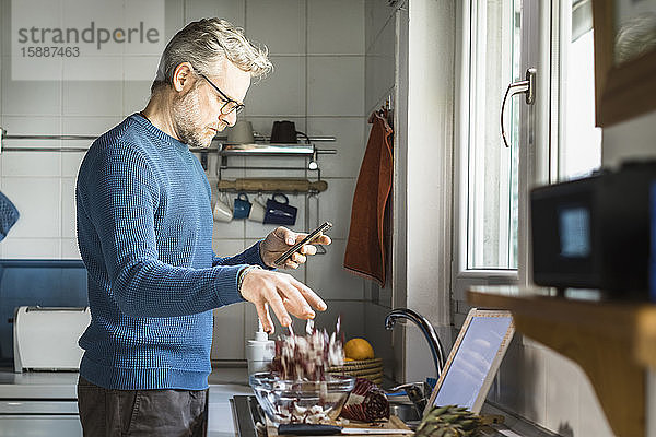 Reifer Mann steht in seiner Küche und schaut auf sein Smartphone  während er Salat zubereitet