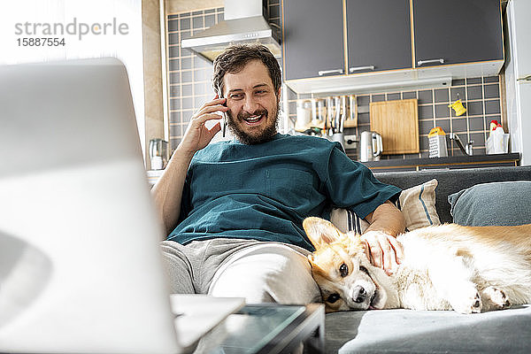 Porträt eines lächelnden Mannes am Telefon  der mit seinem Hund zu Hause auf der Couch sitzt