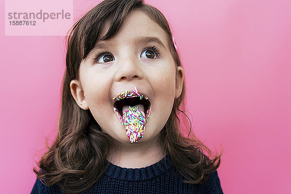 Porträt eines glücklichen kleinen Mädchens mit Zuckergranulat auf Lippen und Zunge vor rosa Hintergrund