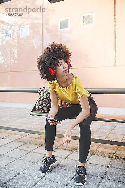 Junge Frau mit Afrofrisur  die an einer Bushaltestelle in der Stadt mit Kopfhörern Musik hört