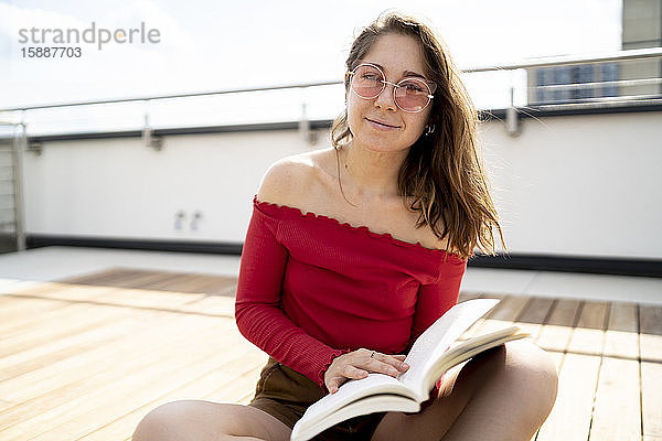 Porträt einer lächelnden jungen Frau beim Lesen eines Buches auf dem Dach