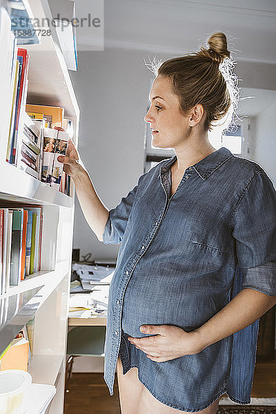Schwangere Frau steht zu Hause am Bücherregal