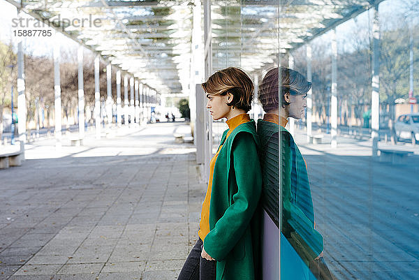 Bildnis einer jungen Frau in grünem Mantel  die sich mit ihrem Spiegelbild an eine bunte Glaswand lehnt
