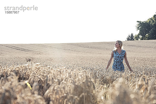 Blonde Frau steht bei Sonnenuntergang in einem Weizenfeld und schaut in die Ferne