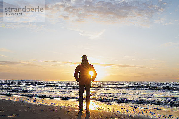 Rückansicht einer Frau in voller Länge  die am Ufer steht  während sie bei Sonnenuntergang das Meer gegen den Himmel betrachtet  Nordseeküste  Flandern  Belgien