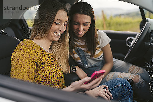 Zwei glückliche junge Frauen auf einer Autoreise  die sich ein Handy teilen