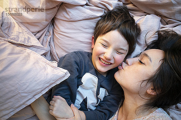Porträt eines lachenden kleinen Jungen  der zu Hause auf dem Bett liegt und von seiner Mutter geküsst wird