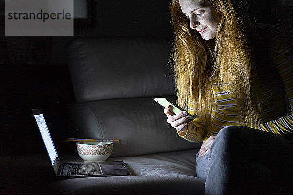 Junge Frau sitzt zu Hause auf der Couch und benutzt Laptop und Smartphone