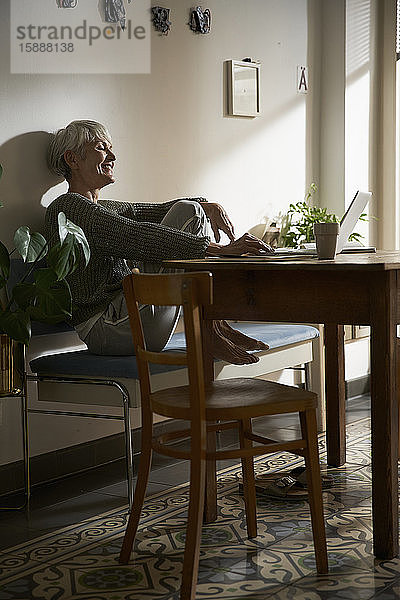 Glückliche ältere Frau sitzt auf Bank in ihrer Küche und benutzt Laptop