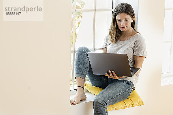 Junge brünette Frau sitzt zu Hause auf der Fensterbank und hält ihren Laptop und arbeitet im Home-Office
