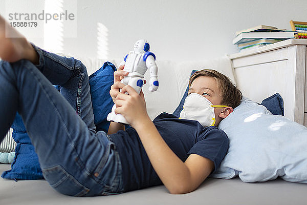 Junge mit Maske spielt während der Koronakrise mit seinem neuen  einzigen Freund  einem Roboter  in seinem Zimmer