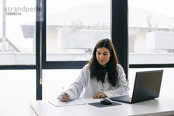 Arzt sitzt am Schreibtisch in medizinischer Praxis und macht Notizen