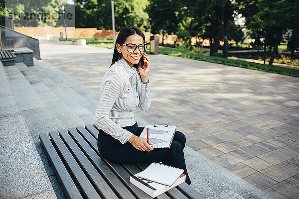 Lächelnde Geschäftsfrau ruft an und sitzt auf einer Bank in einem Park