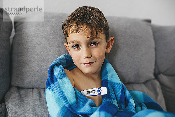 Porträt eines kranken Jungen auf einer Couch sitzend mit Digitalthermometer
