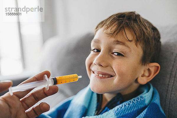 Porträt eines grinsenden kranken Jungen  der auf der Couch sitzt  während seine Mutter ihm Medikamente gibt