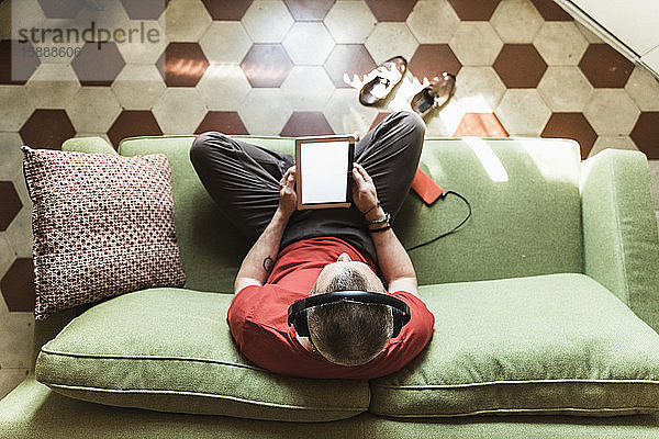 Reifer Mann sitzt im Wohnzimmer auf dem Sofa und benutzt ein Tablett  hört Musik