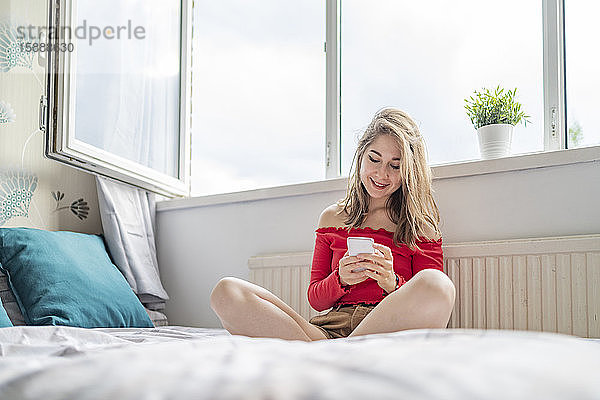 Lächelnde junge Frau sitzt zu Hause auf dem Bett und benutzt ihr Handy