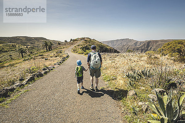 Rückenansicht von Vater und kleinem Sohn mit Rucksäcken  die Hand in Hand auf der Landstraße gehen  La Gomera  Kanarische Inseln  Spanien