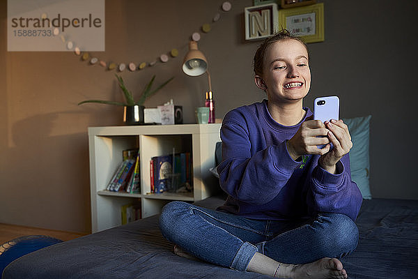 Porträt eines grinsenden Mädchens  das auf dem Bett sitzt und sich mit dem Smartphone selbstständig macht