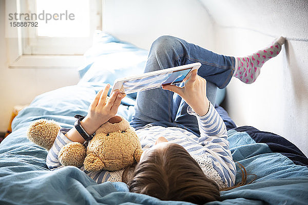 Kleines Mädchen liegt mit Teddybär auf dem Bett und benutzt digitales Tablett