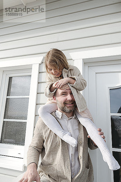 Porträt eines Vaters mit glücklicher Tochter auf den Schultern vor ihrem Haus
