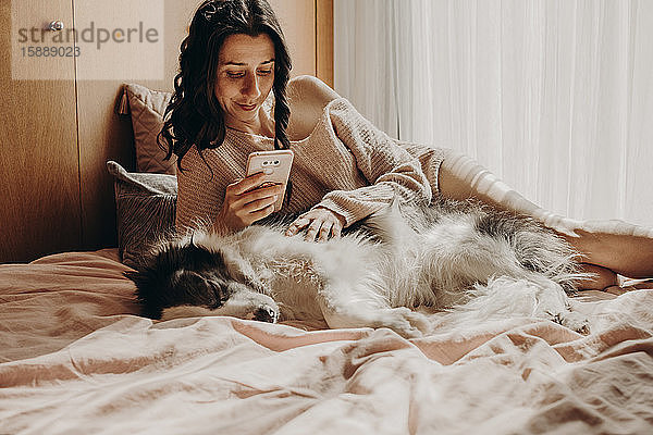 Porträt einer Frau  die sich mit ihrem Hund im Bett entspannt und auf ein Smartphone schaut