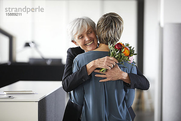 Glückliche ältere Geschäftsfrau mit Blumenstrauss umarmt Amtskollegin