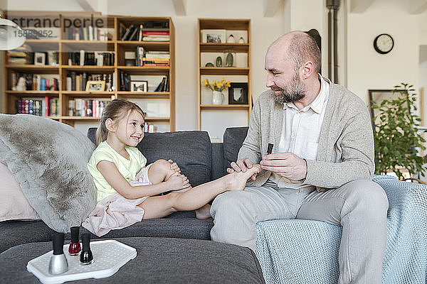 Vater lackiert die Zehennägel seiner Tochter mit rotem Nagellack auf der Couch im Wohnzimmer