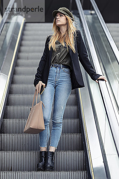 Porträt einer blonden jungen Frau mit Tasche und Smartphone auf einer Rolltreppe stehend