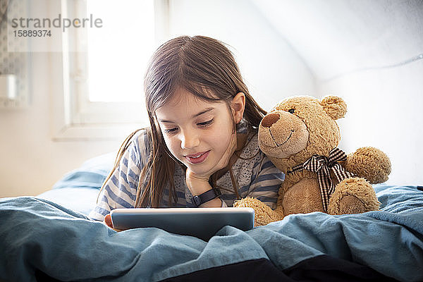 Porträt eines lächelnden Mädchens mit Teddybär auf dem Bett liegend mit digitalem Tablett