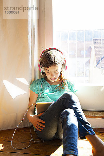 Mädchen sitzt zu Hause auf dem Boden und benutzt Kopfhörer und digitales Tablet