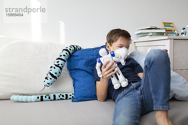 Junge mit Maske spielt während der Koronakrise in seinem Zimmer mit seinem neuen  einzigen Freund  einem Roboter und einem Smartphone