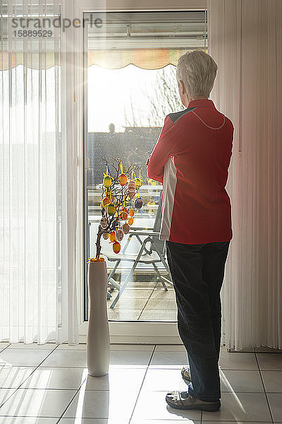Ältere Frau steht auf geschlossenem Balkon und schaut aus dem Fenster